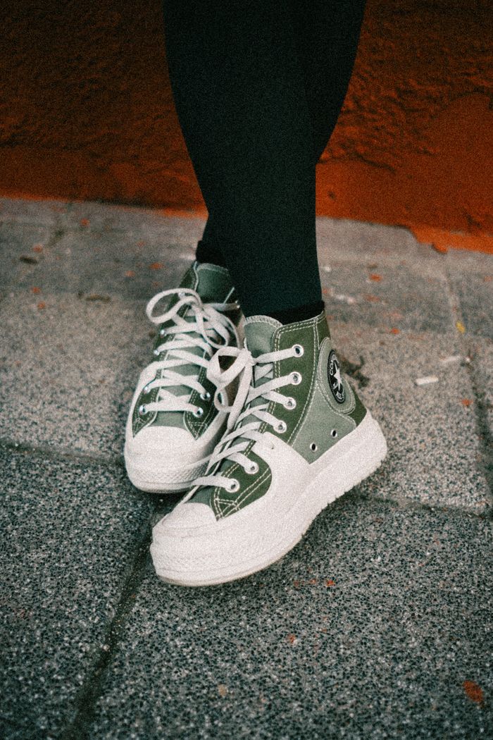 Green sneakers with white laces on pavement, captured outdoors with an orange wall backdrop.